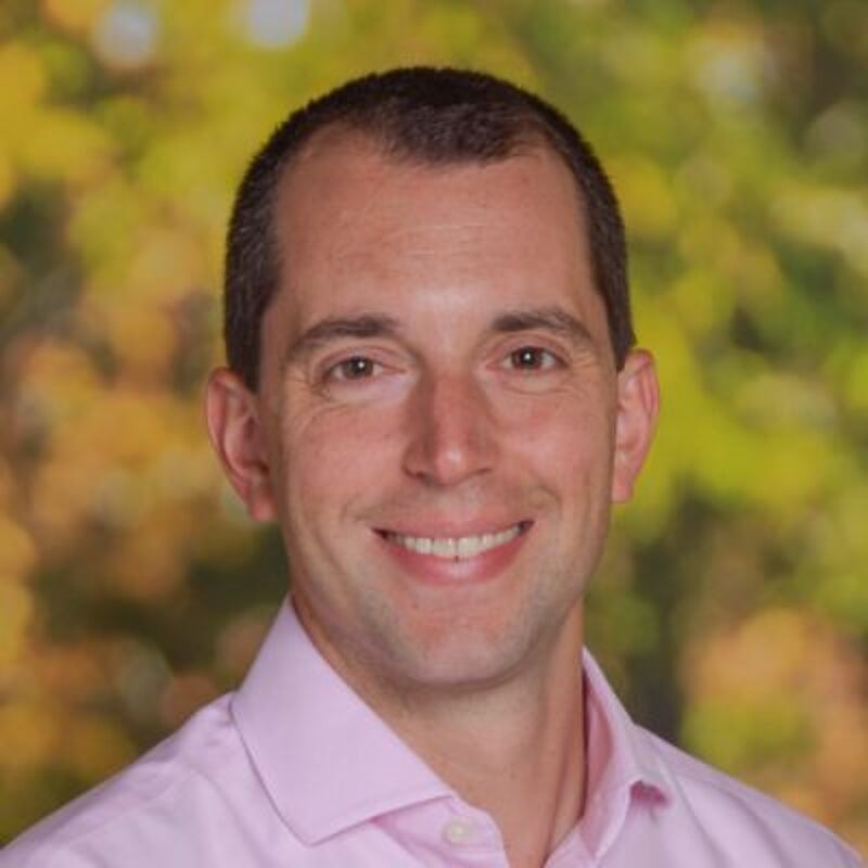The image shows a man with short brown hair and brown eyes. He is wearing a pink shirt and smiling at the camera. The background is blurred, with shades of green and yellow, suggesting an outdoor setting. The man appears to be in his late 30s or early 40s.